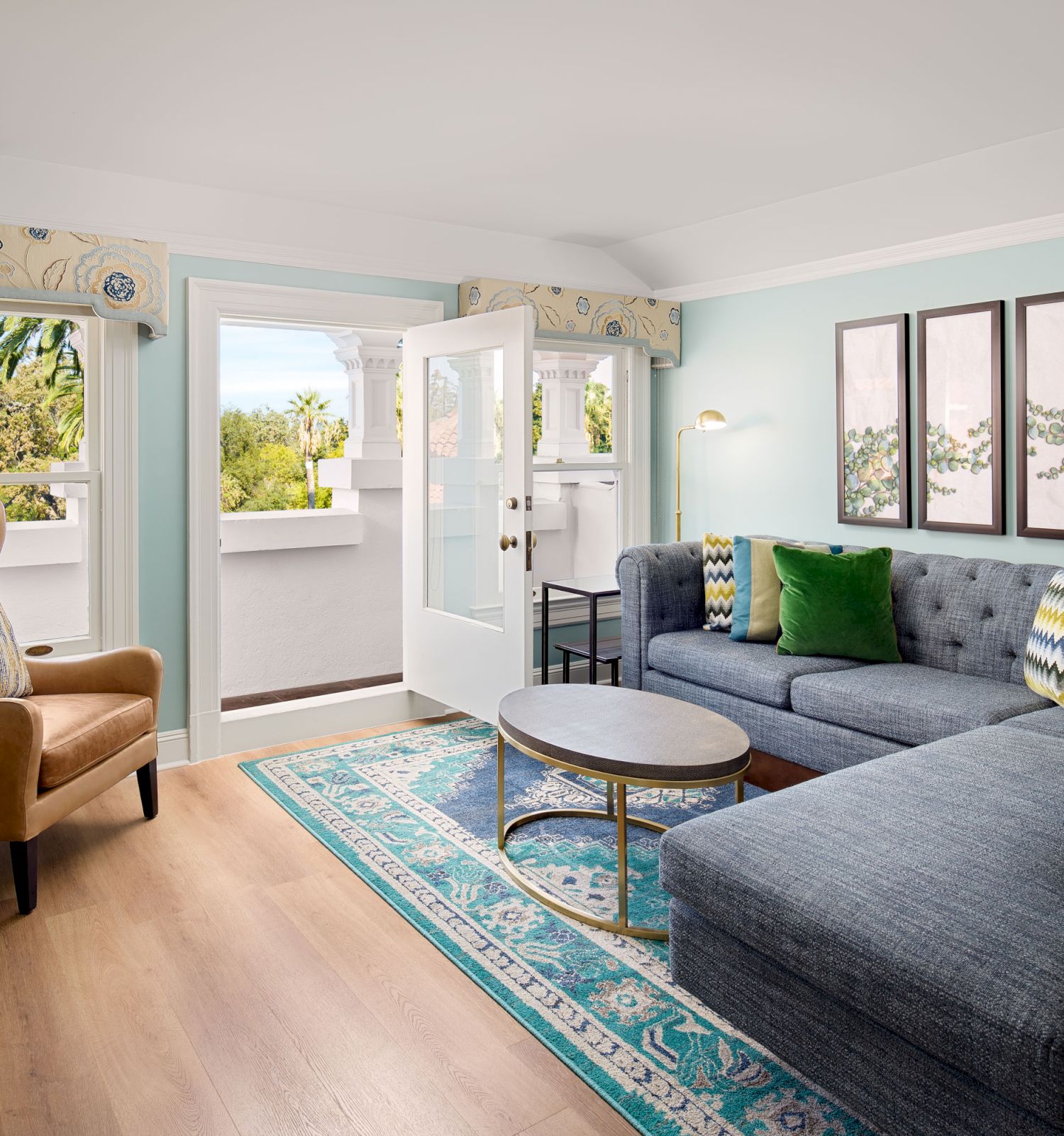 A bright living room with a blue sectional sofa, patterned rug, armchair, round coffee tables, and a door opening to a balcony under soft pastel walls.