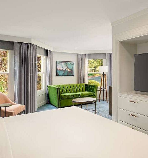A bright hotel suite with a white bed, a green sofa, pink chair, large windows with blue curtains, a dresser with a flat-screen TV, and a cozy seating area.