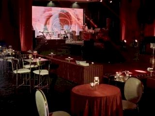 A dim, intimate stage with red lighting, round tables with chairs, and a projection screen behind the performers.