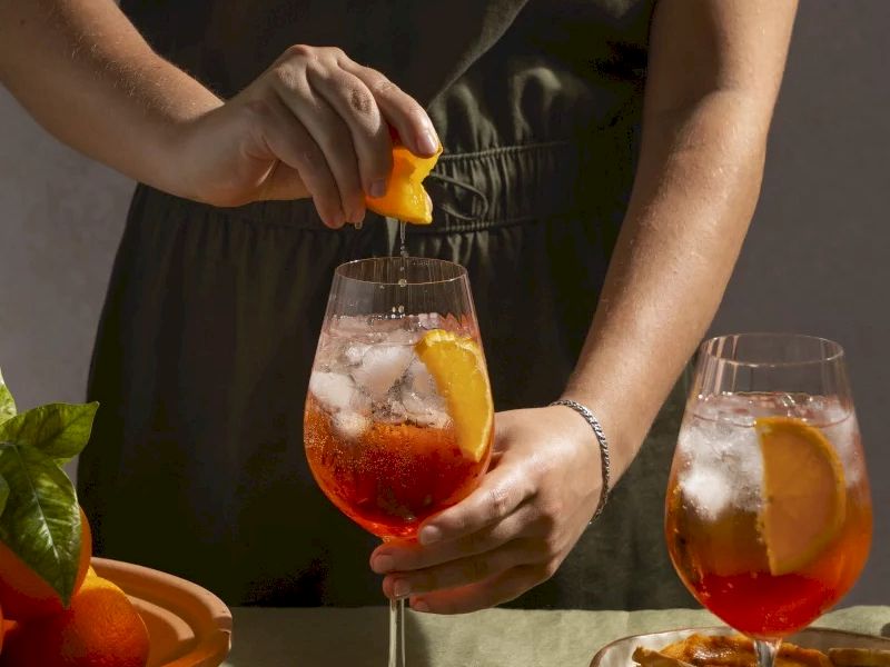 A person&rsquo;s hand is squeezing a lemon over two iced cocktails in stemmed glasses, with fruit and plates on the table.