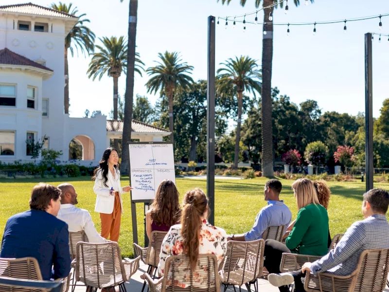 Outdoor wedding or event ceremony setup with chairs facing a speaker podium, string lights, palm trees, and attendees in a sunny garden.