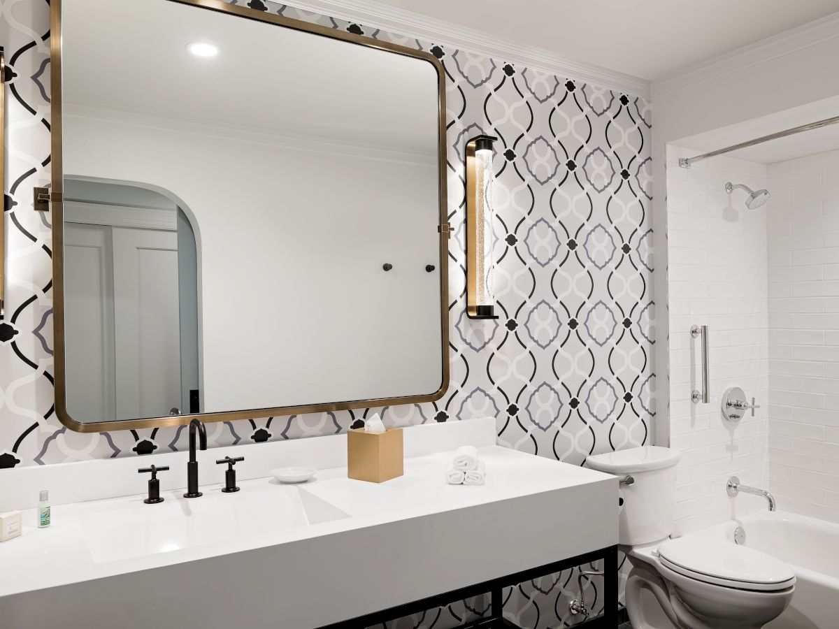 A modern bathroom with a large framed mirror, brass light fixtures, patterned tiles, a white sink, and a toilet.