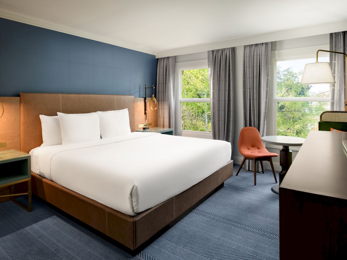 A modern hotel room with a large bed, bedside lamps, gray curtains, and a desk by big windows overlooking green trees.