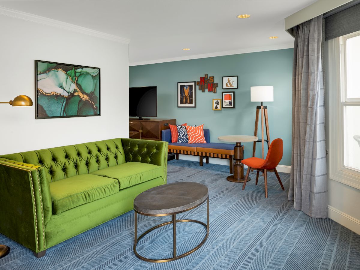 Bright living room with a green tufted sofa, patterned rug, teal accent wall, framed art, and an orange chair by the window.