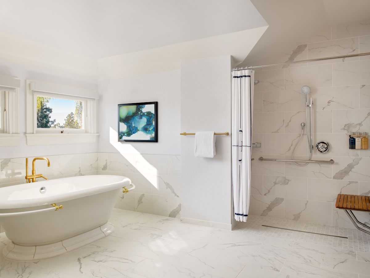 Bright luxury bathroom with marble tiles, a soaking tub, window, framed art, and a glass-enclosed walk-in shower with gold fixtures.