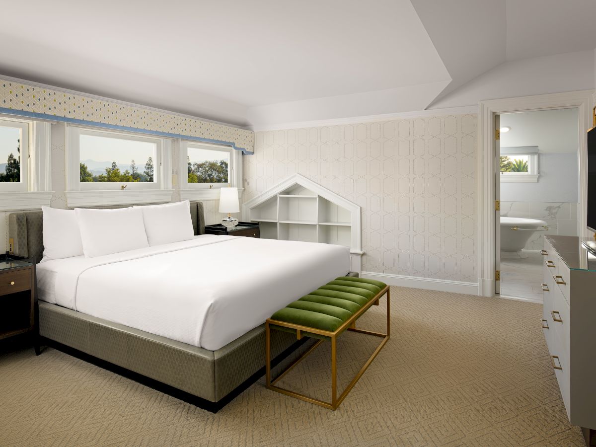 A modern hotel bedroom with a large bed, patterned wallpaper, windows with views, and an open vanity/bath area.