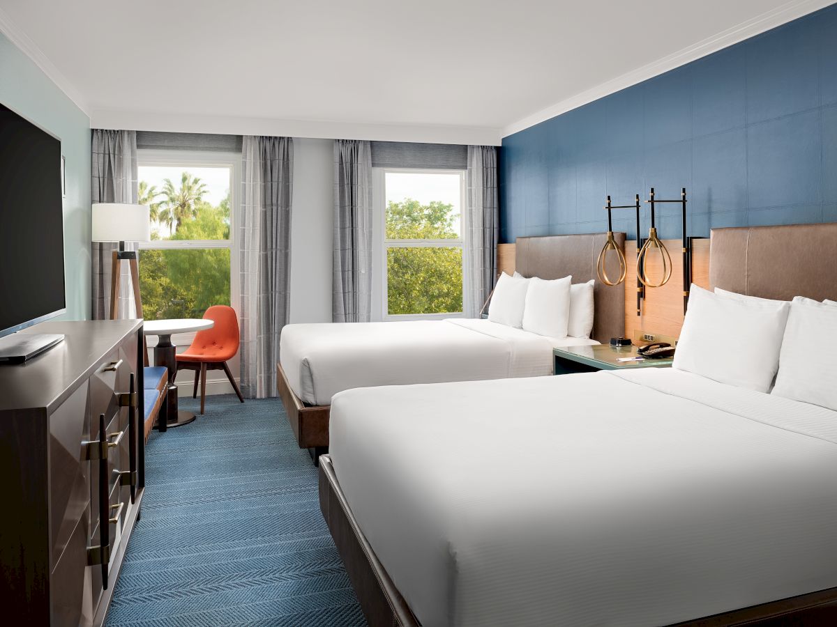 A modern hotel room with two white beds, a blue feature wall, curtained windows, and a small desk near the entrance.