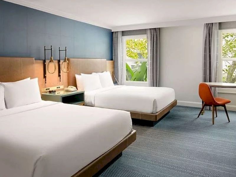 Two neatly made twin beds in a modern hotel room with a blue accent wall, desk chair, and window view, ending with a period.