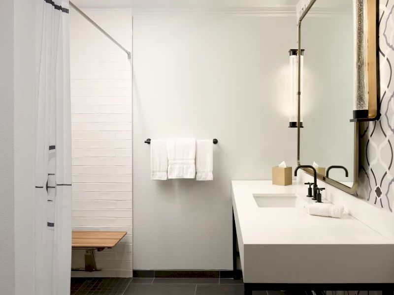 A modern bathroom with a double sink, white vanity, black fixtures, patterned wall, a glass shower door, and a wooden bench in a light, airy space.