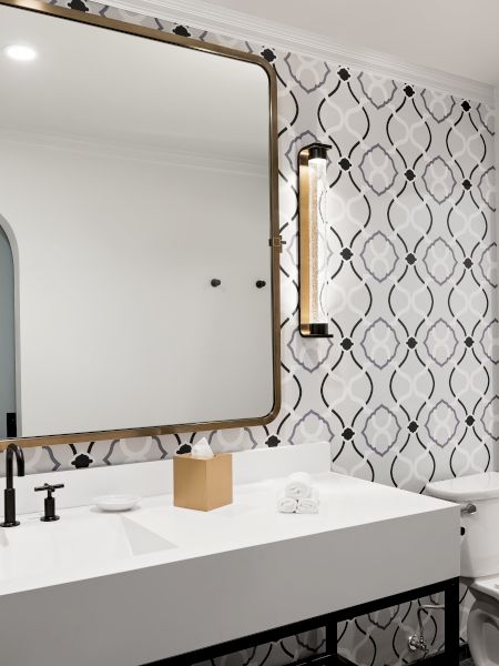 A modern bathroom with a large framed mirror, gold sconces, patterned tile/wallpaper, a white vanity, and a toilet in view.