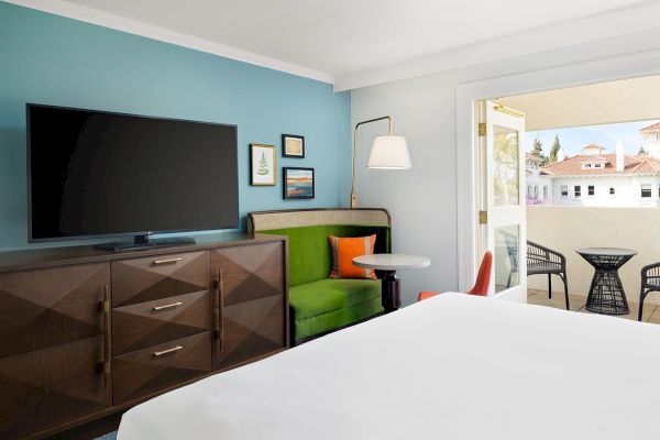 A modern bedroom with a wall TV, dark dresser, green bench seating, a floor lamp, and a window opening to a patio with chairs.