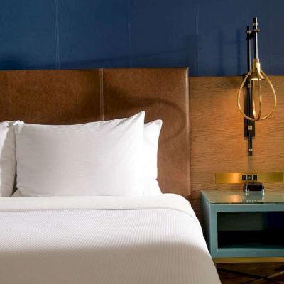 A neatly made bed sits beside a wooden nightstand with a glass-topped lamp in a modern blue-walled room.