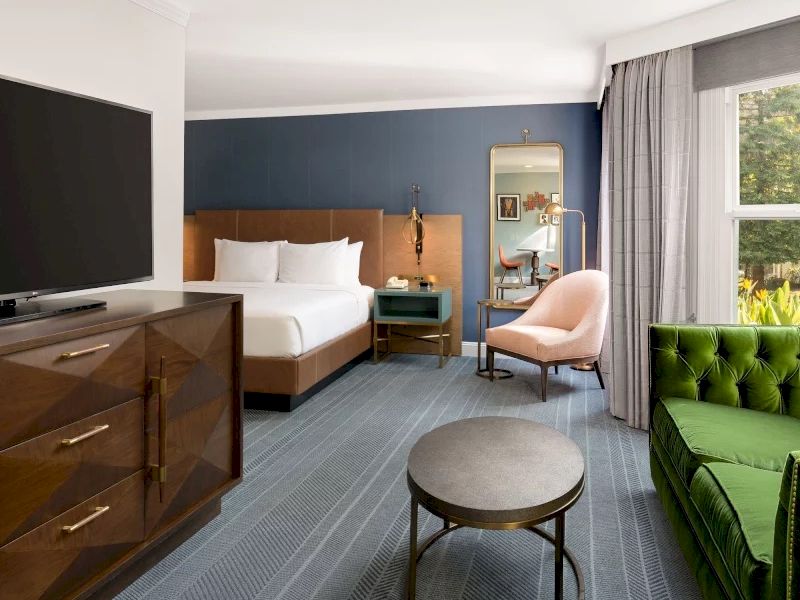 A hotel room with a bed, blue accent wall, dresser, TV, chair, round coffee table, green tufted sofa, and large window with curtains.