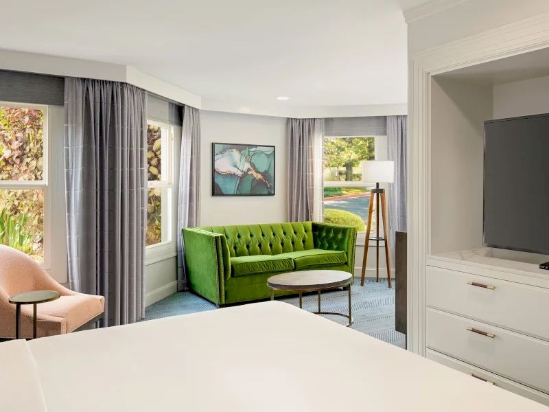 A bright modern living room/office with a green tufted sofa, curtains, framed art, and a large TV mounted on white cabinetry.
