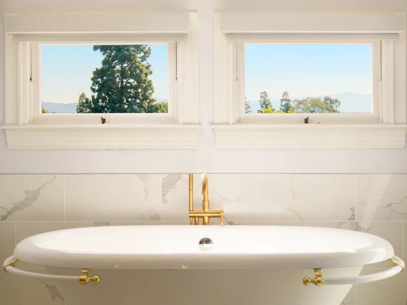 A bright, airy bathroom with a freestanding tub and gold fixtures, framed by two small windows overlooking trees and a clear sky.