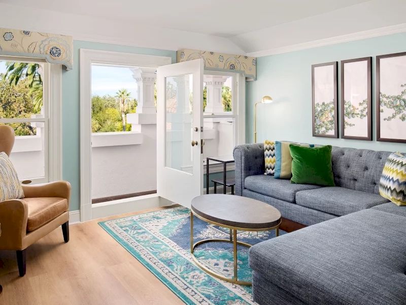 A bright, airy living room with light blue walls, a large blue sectional sofa, colorful cushions, a round coffee table, and a patterned turquoise rug; sunlight streams in through open doors to a balcony.