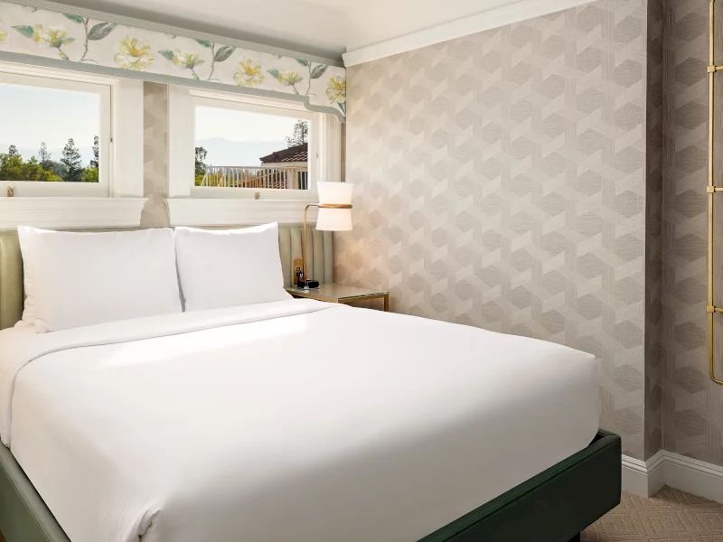 A neat hotel room with a king bed, white linens, wall pattern, soft lighting, and a window view. All clean and cozy.