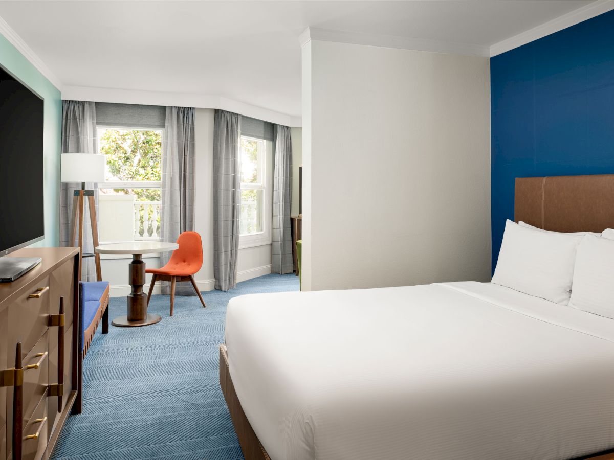 A modern hotel room with a white bed, blue accent wall, desk and chair by large windows with gray curtains.