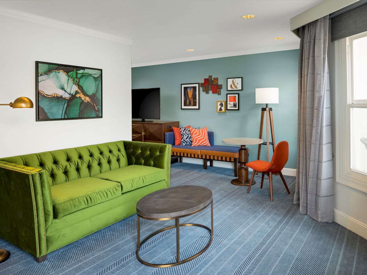 A modern lounge with green sofa, blue accent wall, patterned rug, framed art, a floor lamp, and chairs by the window.