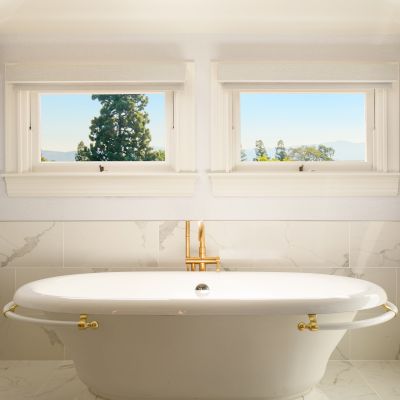 A bright modern bathroom with a white soaking tub, marble walls, two windows, and a framed wall TV.