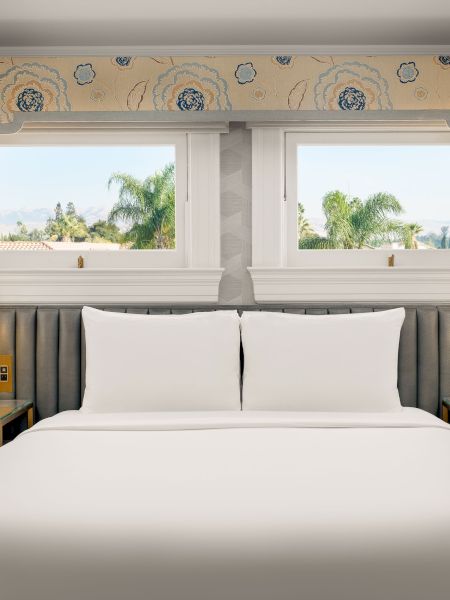 A modern bedroom with a large bed, gold lamps, blue-gray curtains, and two windows showing palm trees and hills outside.