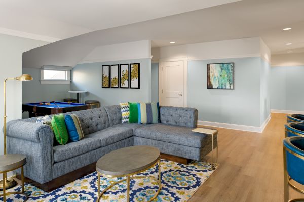 A bright living room with a gray sectional sofa, colorful pillows and rug, framed art, and a pool table in the back.