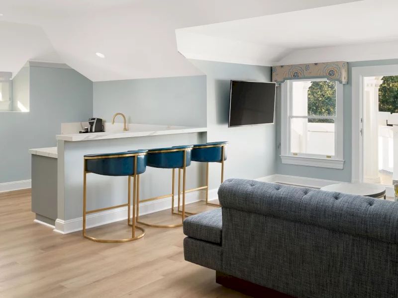 A bright, modern living area with a light wood floor, blue kitchen nook, bar seating, a gray sofa, and a wall-mounted TV near windows, open layout.