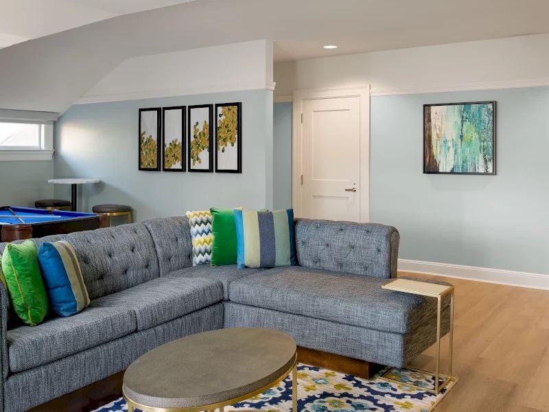 Cozy living room with a light blue wall, gray sectional, round coffee table, colorful patterned rug, framed art, and natural light. All set for relaxation.