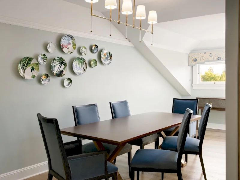 A modern dining room with a dark wood table and eight blue-chaired seating, a light chandelier, and decorative plates on a pale green wall.