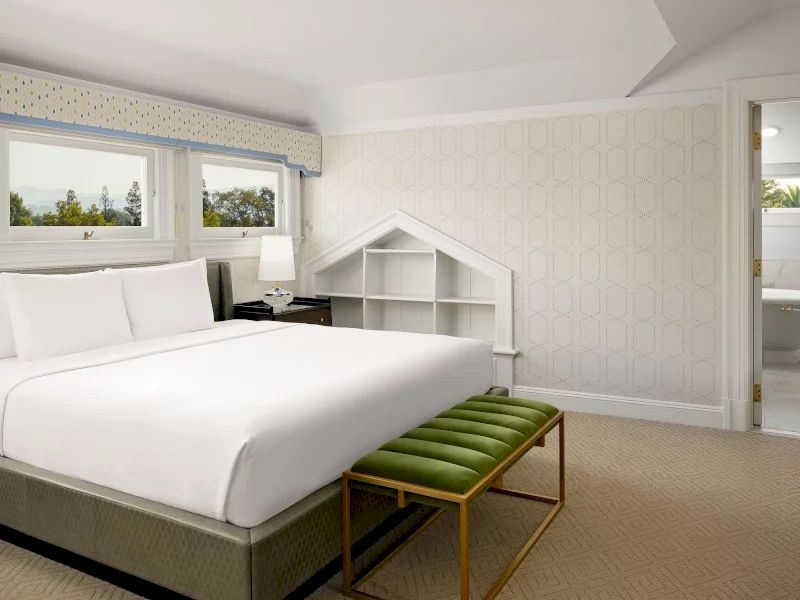 A bright hotel bedroom with a large bed, white linens, a small desk, a shelf unit by the wall, and a green upholstered bench at the foot, neutral tones everywhere.