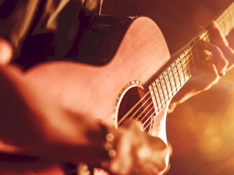 A close-up of a person playing an acoustic guitar on stage, warm lighting glowing around the instrument.