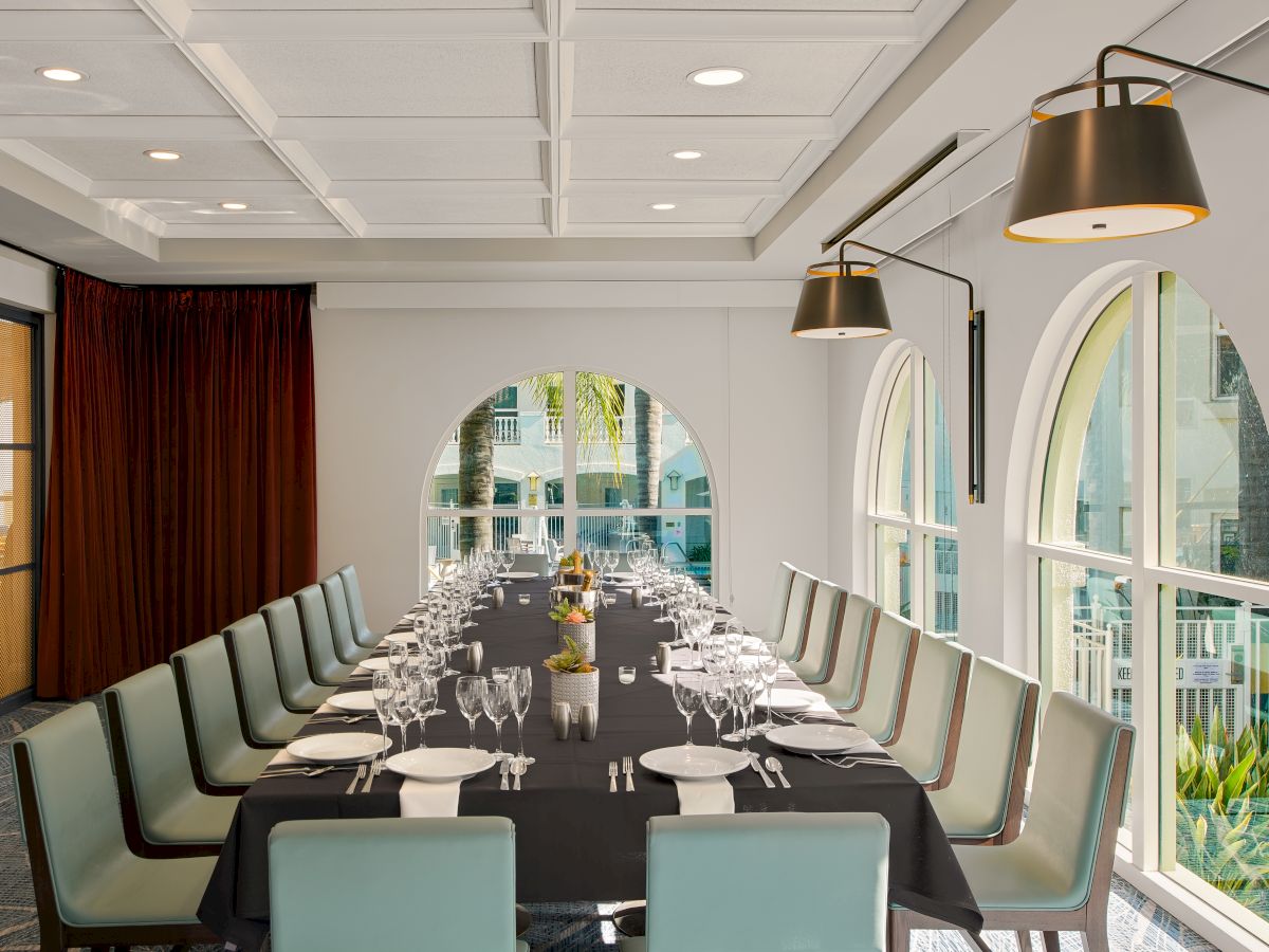 A modern banquet dining room with a set table, crystal glassware, arched windows, and warm pendant lamps.