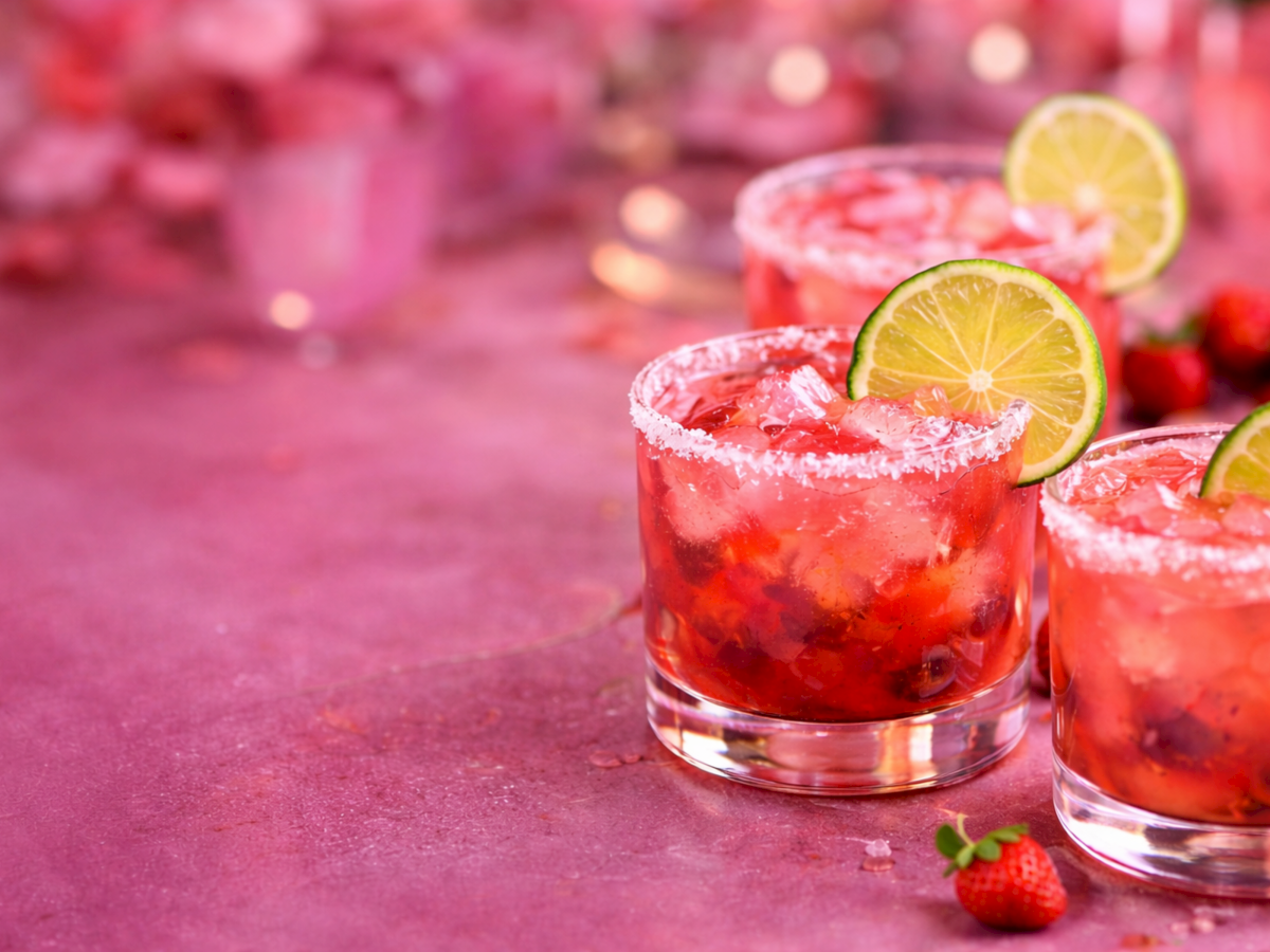 Bright red berry cocktails in glasses with crushed ice, garnished with fresh lime/lemon slices on a pink backdrop.