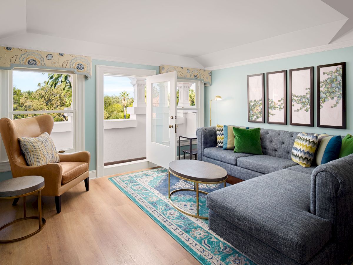 A bright living room with a blue sectional sofa, patterned rug, accent chairs, framed art, and large open doorways to the balcony.