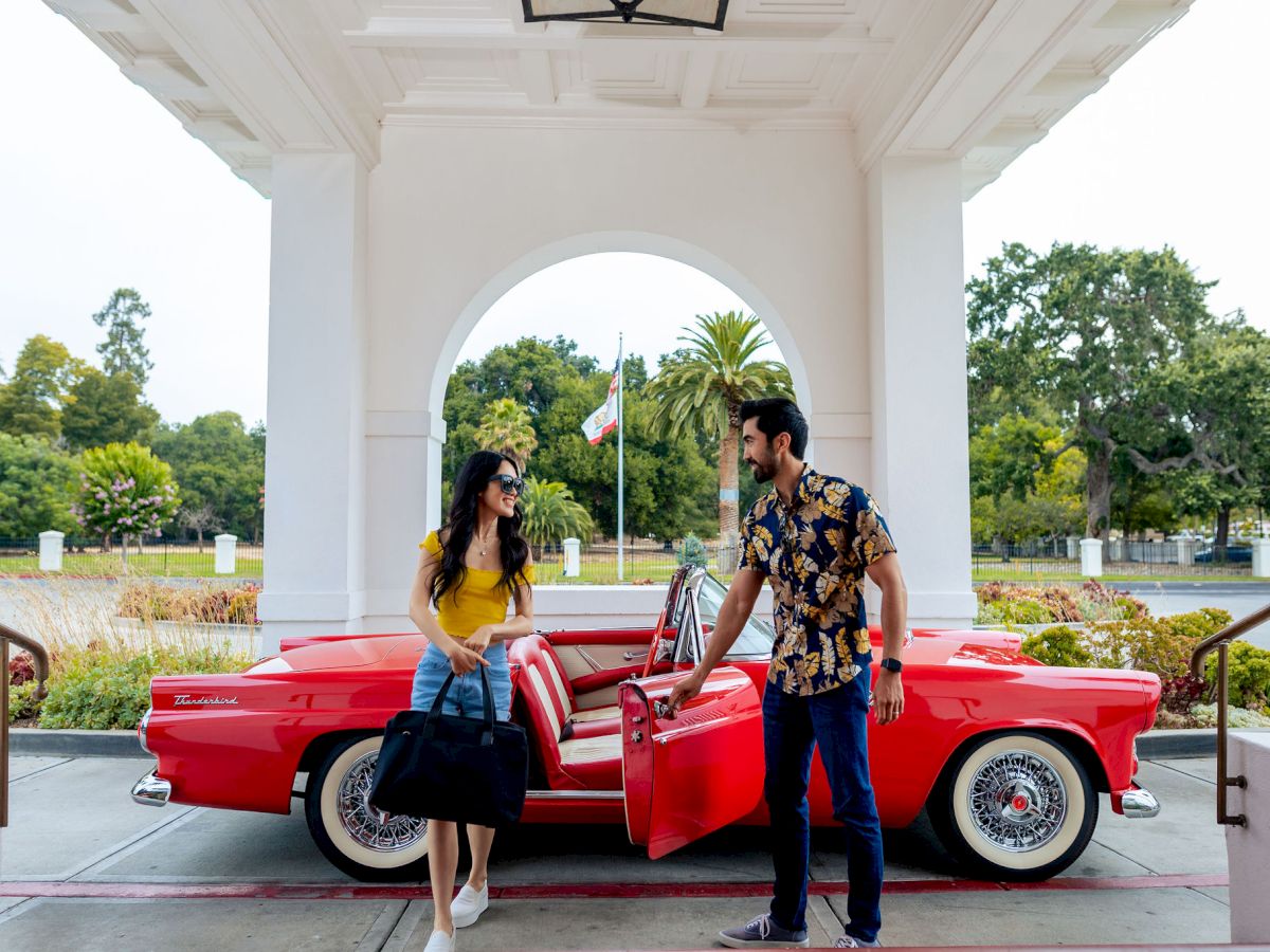 A couple stands by a bright red classic convertible under a white archway, smiling and posing outdoors among lush greenery.