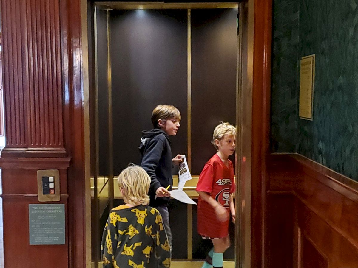Four kids stand in a doorway inside a building, watching and holding papers as they look around.