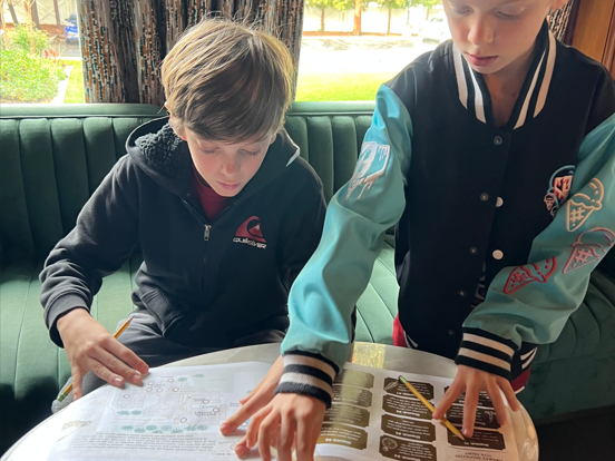 Two kids in jackets sit on a green booth, pointing at a printed map/guide while reading it together outside.