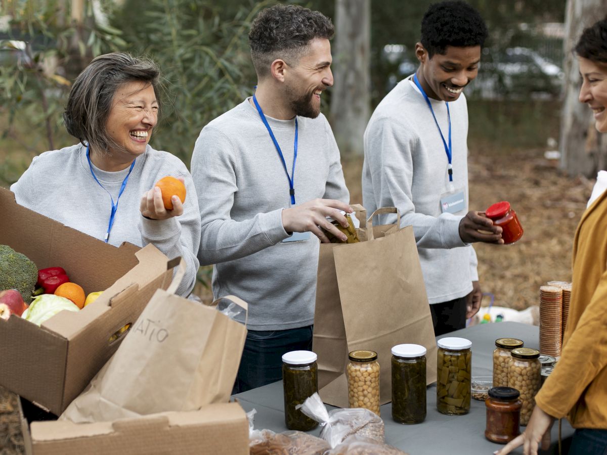 Volunteers pack food boxes with fruits, groceries, and jars outdoors, smiling as they assemble meal supplies for others.