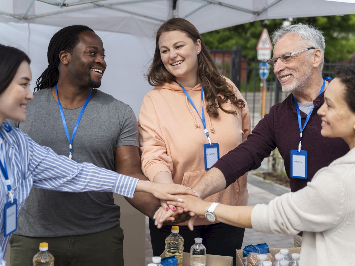 A group of volunteers under a tent smile and hold hands together, suggesting teamwork and community service.
