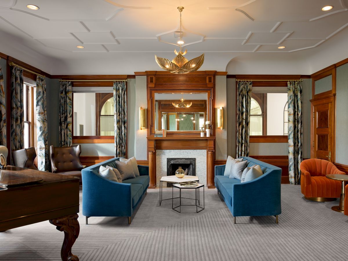 An elegant lounge with blue sofas, a fireplace, ornate woodwork, and a gold chandelier overhead, framed by arched windows.