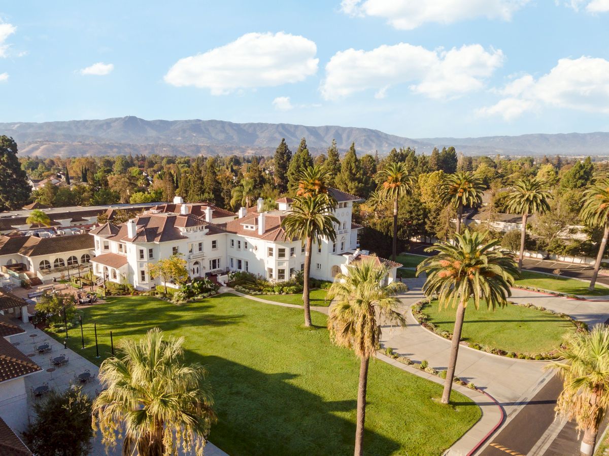 Aerial view of a white villa/hotel with tiled roofs, manicured lawns, palm trees, and a distant mountain range under clouds.