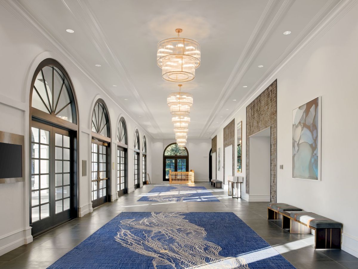 Bright hallway with arched windows, gold chandeliers, framed art, and a large blue patterned rug on polished floors.