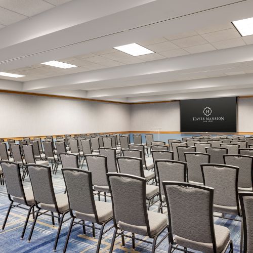 A conference room with many gray upholstered chairs arranged in rows facing a stage and a large screen that reads &ldquo;LIF&hellip;.&rdquo;.