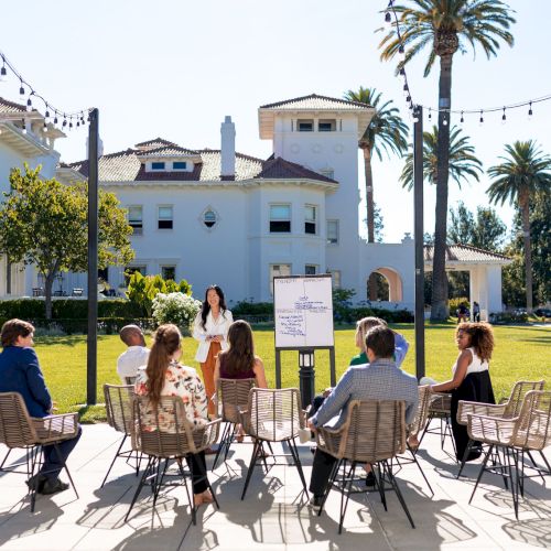 A group of people sits in a semicircle on metal chairs outside a white villa, listening to a speaker with a signboard, sunny day.