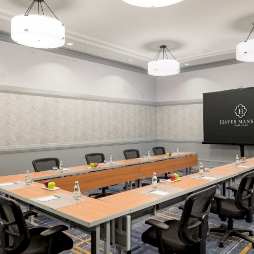 A modern conference room with a U-shaped table, black chairs, water bottles, and a large screen displaying &ldquo;BETTER MANSION&rdquo; in a formal setting.