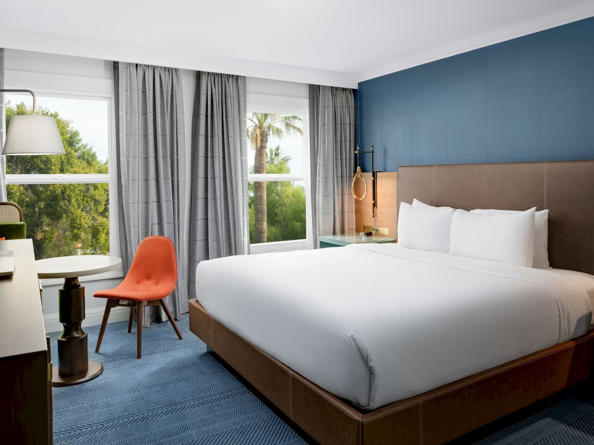 A modern hotel bedroom with a large bed, gray curtains, blue accent wall, and a desk chair by big windows.