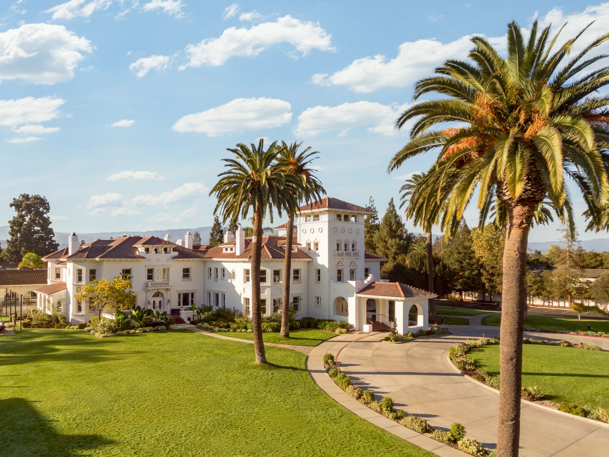 A white resort mansion with red-tile roofs, manicured lawn, and tall palm trees under a bright blue sky.