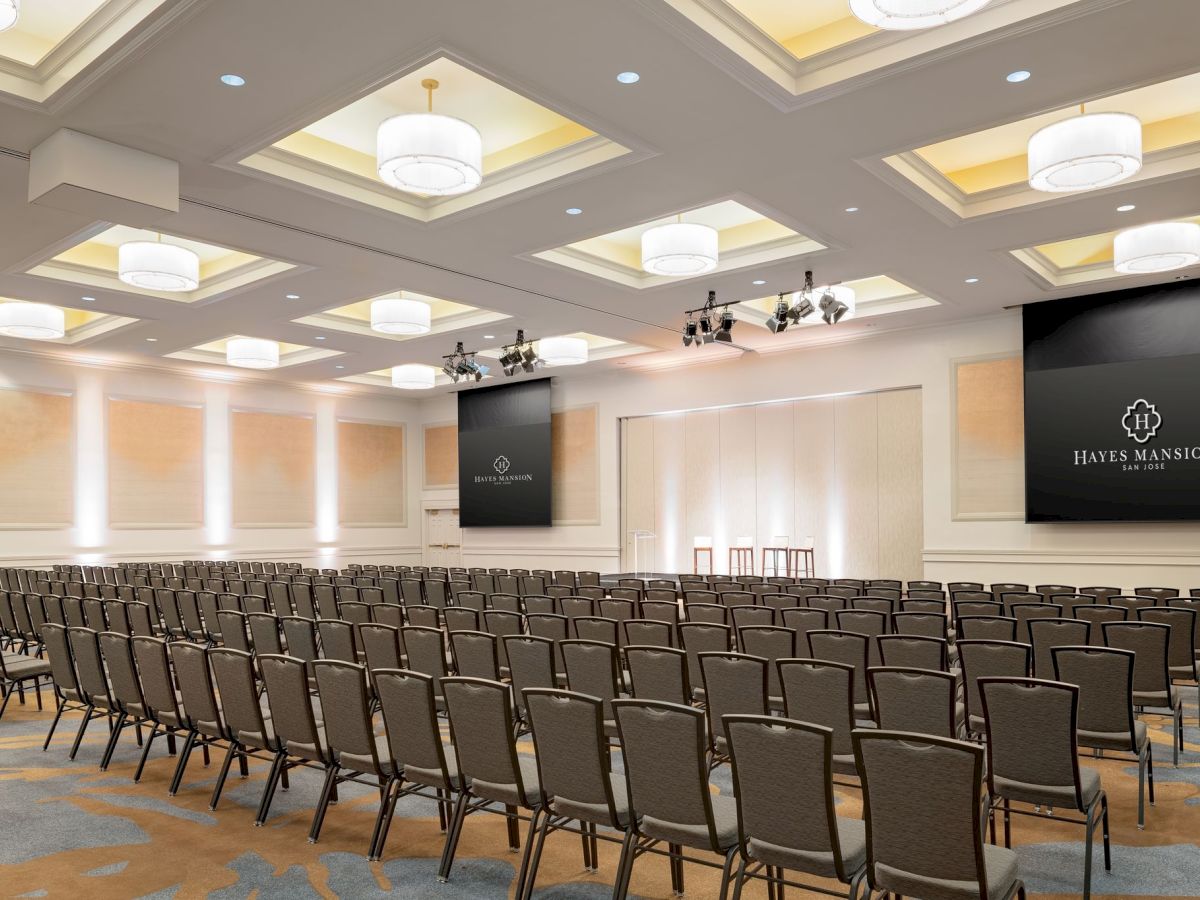 A modern conference hall with rows of chairs, two large screens, chandeliers, and a stage&mdash;Hayes Mansion San Jose branding.