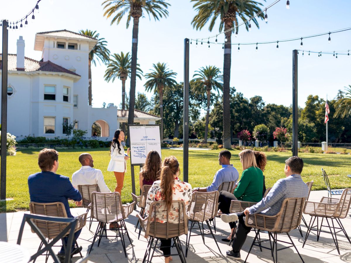 An outdoor group class: a teacher presents a whiteboard while students sit at tables on a sunny lawn with palm trees.