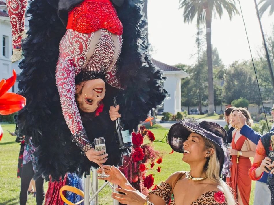 A masked performer in black feathers and red patterned costume cheers with guests at an outdoor party with drinks and flowers.